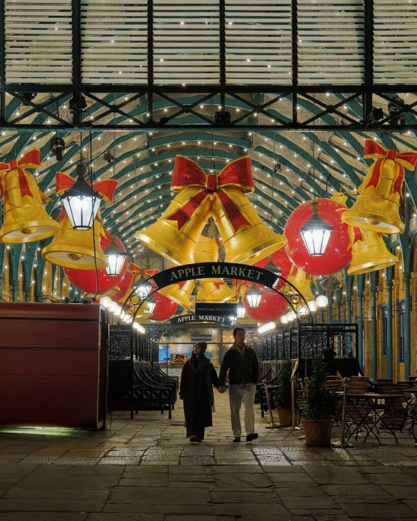 A couple walks through the festive Apple Market in Covent Garden, London, adorned with holiday decorations.